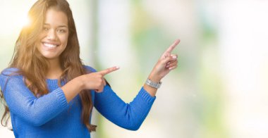 Gülümseyen ve iki elleri ve parmakları ile tarafına işaret kamera bakarak izole arka plan üzerinde mavi kazak giyen genç güzel esmer kadın.