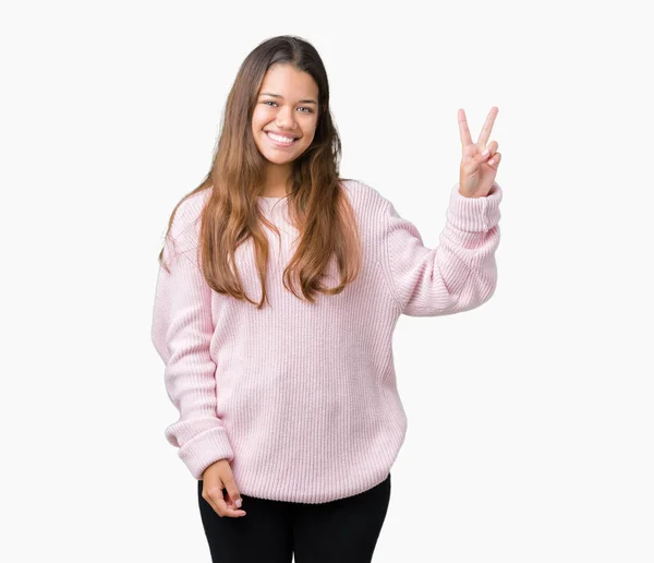 年轻美丽的黑发女人穿着粉红色的冬季毛衣在孤立的背景微笑着与幸福的脸眨眼在相机做胜利的标志 — 图库照片