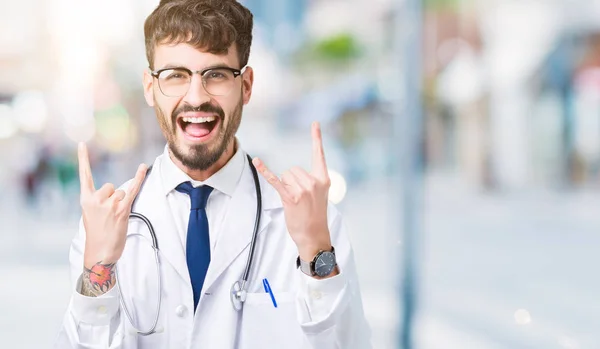 Young doctor man wearing hospital coat over isolated background shouting with crazy expression doing rock symbol with hands up. Music star. Heavy concept.