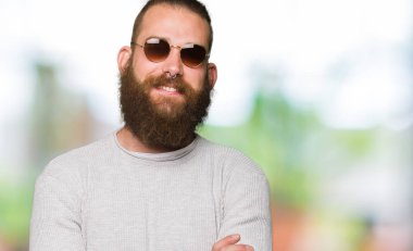 Kameraya bakarak çapraz silah ile gülümseyen güneş gözlüğü mutlu yüz giyen genç hipster adam. Olumlu bir kişi.