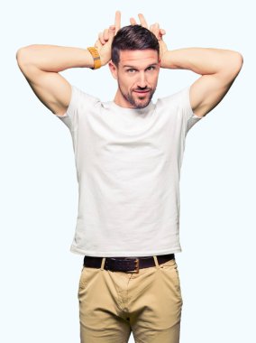 Yakışıklı adam neşeli gülümseyen komik ve çılgın baş parmaklar ile tavşan kulakları poz casual beyaz t-shirt giyen