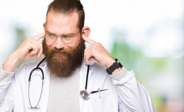 Yüksek sesle müzik gürültü rahatsız ifade parmaklarıyla kulaklarıyla kapsayan bir tıbbi palto giyiyor sakallı genç sarışın doktor adam. Sağır kavramı.