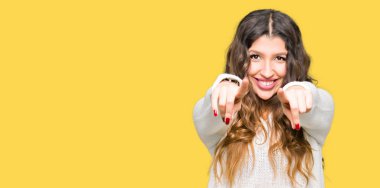 Beyaz kazak giyen genç güzel kadın seni ve kameraya gülümseyen pozitif ve neşeli parmaklarıyla işaret