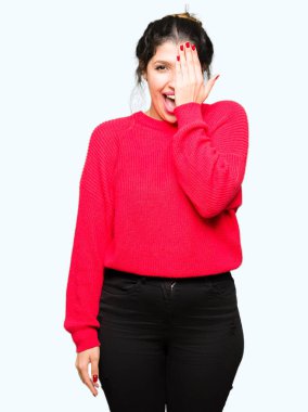 Kırmızı kazak ve el yüz ve sürpriz duygu kendinden emin gülümseme ile tek gözüyle kapsayan topuz giyen genç güzel kadın.