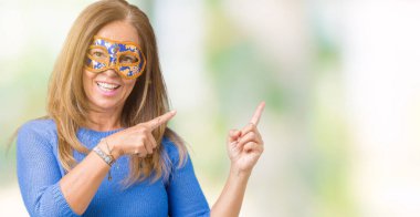 Gülümseyen ve iki elleri ve parmakları ile tarafına işaret kamera bakarak izole arka plan üzerinde Venedik karnaval maskesi giyen güzel orta yaş kadın.