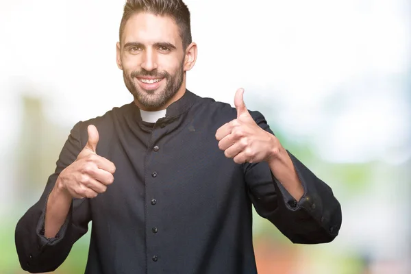 Young Christian priest over isolated background success sign doing positive gesture with hand, thumbs up smiling and happy. Looking at the camera with cheerful expression, winner gesture.