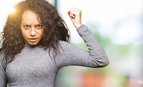 Young beautiful girl with curly hair angry and mad raising fist frustrated and furious while shouting with anger. Rage and aggressive concept.