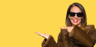 Güzel orta yaş zarif kadın gözlük ve vizon ceket giyiyor hayran ve kameraya gülümseyen el ile sunmak ve parmak ile işaret.