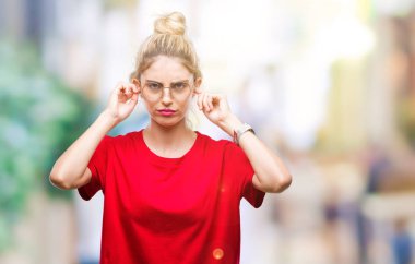 Yüksek sesle müzik gürültü rahatsız ifade parmaklarıyla kulaklarıyla kapsayan izole arka plan üzerinde kırmızı t-shirt ve gözlük giyen genç güzel sarışın kadın. Sağır kavramı.