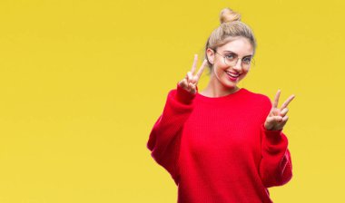 Zafer işareti yapıyor parmak gösterilen kamera seyir gülümseyen izole arka plan üzerinde kırmızı kazak ve gözlük giyen genç güzel sarışın kadın. İki numaralı.