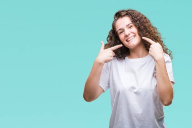 Güzel esmer kıvırcık saçlı Genç kız kendinden emin gösterilen ve işaret parmakları diş ve ağız ile gülümseyen izole arka plan üzerinde rahat t-shirt giyiyor. Sağlık kavramı.
