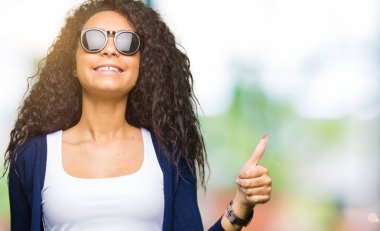 Genç güzel kız kıvırcık saçlı hareketi el ile mutlu yaşasın yapıyor moda güneş gözlüğü takıyor. Kameraya bakarak başarı gösteren ile ifade onaylama.