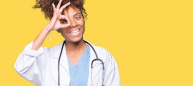 Genç Afro-Amerikan doktor kadın gülümsüyor, parmakları ile mutlu yüz üzerinden seyir göz el ile ok jest yapıyor izole arka plan.