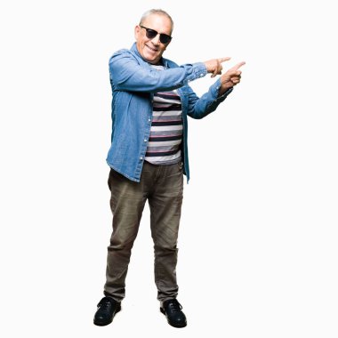Yakışıklı kıdemli adam giyen denim ceket ve gülümseyen ve iki elleri ve parmakları ile tarafına işaret kamera bakarak güneş gözlüğü.