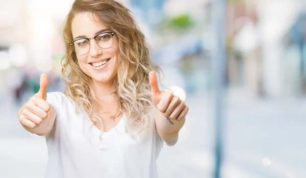 Beautiful young blonde woman wearing glasses over isolated background approving doing positive gesture with hand, thumbs up smiling and happy for success. Looking at the camera, winner gesture.