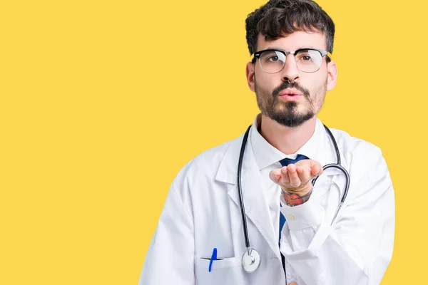 Young doctor man wearing hospital coat over isolated background looking at the camera blowing a kiss with hand on air being lovely and sexy. Love expression.