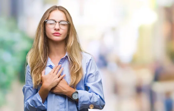 Young beautiful blonde business woman wearing glasses over isolated background smiling with hands on chest with closed eyes and grateful gesture on face. Health concept.
