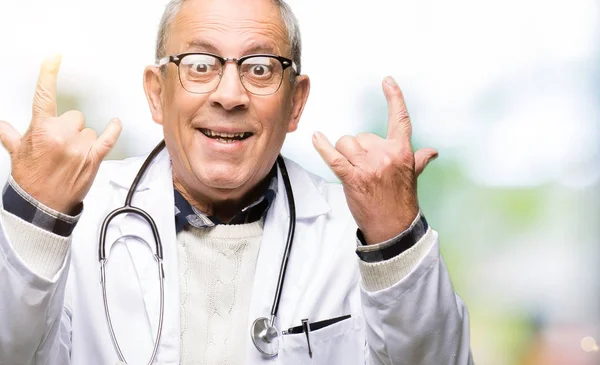 Handsome senior doctor man wearing medical coat shouting with crazy expression doing rock symbol with hands up. Music star. Heavy concept.