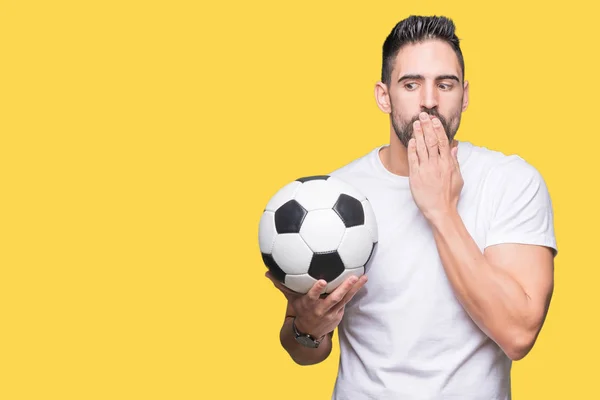 Young man holding soccer football ball over isolated background cover mouth with hand shocked with shame for mistake, expression of fear, scared in silence, secret concept