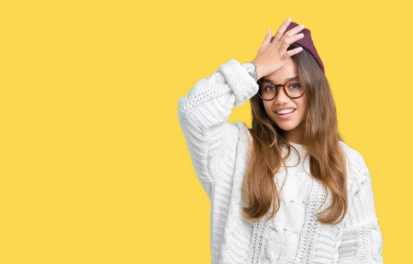 Young beautiful brunette hipster woman wearing glasses and winter hat over isolated background surprised with hand on head for mistake, remember error. Forgot, bad memory concept.