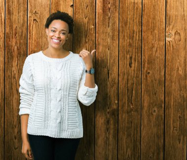 Seyir ve başparmak yanında yukarıyı mutlu yüz ile gülümseyen izole arka plan üzerinde kazak giyen güzel genç Afro-Amerikan kadın.