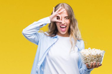 Mutlu yüz el parmaklarının arasından seyir göz üzerinde ok işaretiyle yapıyor gülümseyen izole arka plan üzerinde patlamış mısır yemek güzel sarışın genç kadın