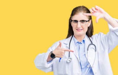 Orta yaş olgun doktor kadın elle yapma çerçeve ve parmakları ile mutlu yüz gülümseyen izole arka plan üzerinde tıbbi bir palto giyiyor. Yaratıcılık ve Fotoğrafçılık kavramı.