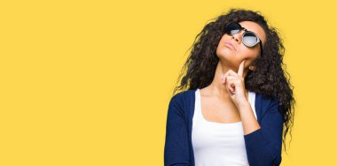 Kıvırcık saçlı Genç güzel kız, soru, dalgın ifade hakkında çene düşünme el ile moda güneş gözlüğü giyiyor. Düşünceli bir yüz ile gülümsüyor. Şüphe kavramı.