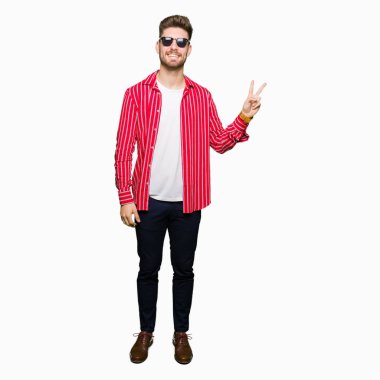 Genç yakışıklı adam mutlu yüz zafer işareti yaparken kameraya göz kırpıyor ile gülümseyen güneş gözlüğü takıyor. İki numaralı.