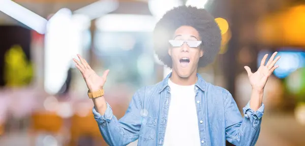 Young african american man with afro hair wearing thug life glasses celebrating crazy and amazed for success with arms raised and open eyes screaming excited. Winner concept