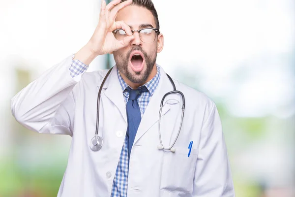 Handsome young doctor man over isolated background doing ok gesture shocked with surprised face, eye looking through fingers. Unbelieving expression.