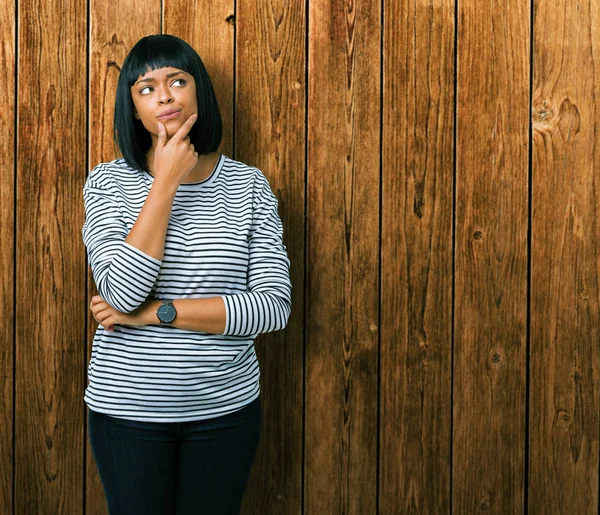Beautiful young african american woman wearing stripes sweater over isolated background with hand on chin thinking about question, pensive expression. Smiling with thoughtful face. Doubt concept.