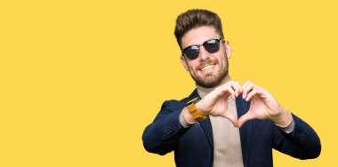 Genç yakışıklı zarif adam aşık kalp simgesi ve elleriyle şekil gösteren gülümseyen güneş gözlüğü takıyor. Romantik kavramı.