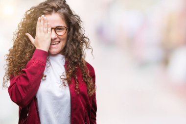 Ceket ve gözlük el yüz ve sürpriz duygu kendinden emin gülümseme ile tek gözüyle kapsayan izole arka plan üzerinde giyen güzel esmer kıvırcık saçlı Genç kız.