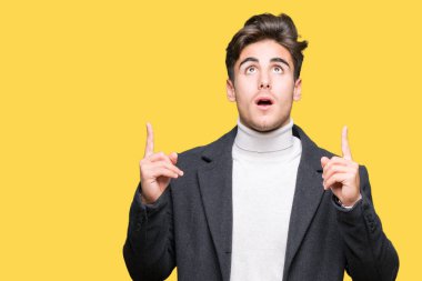 Zarif delikanlı izole arka plan üzerinde kış palto giyiyor hayrete ve yukarıya bakıyor ve parmakları ile işaret ve silah kaldırdı şaşırttı.