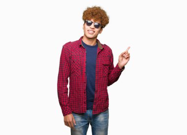 Kameraya bakarak yan el ve parmak ile işaret eden genç yakışıklı adam, yüzünde kocaman bir gülümseme ile güneş gözlüğü takıyor afro saçlı.