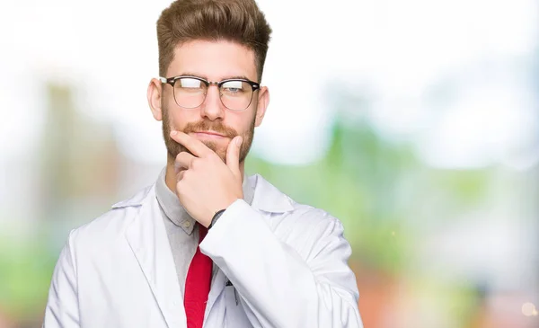 Young handsome scientist man wearing glasses looking confident at the camera with smile with crossed arms and hand raised on chin. Thinking positive.