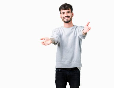 Sarılmak için kollarını açarak gülümseyerek kameraya bakarak izole arka plan üzerinde sweatshirt giyen genç yakışıklı adam. Neşeli ifade kucaklayan mutluluk.