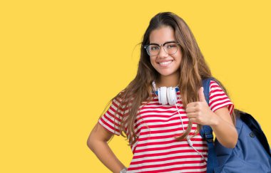 Genç güzel esmer öğrenci kadın hareketi el ile mutlu yaşasın yapıyor izole arka plan üzerinde kulaklık ve sırt çantası giyiyor. Kameraya bakarak başarı gösteren ile ifade onaylama.