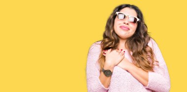 Güneş gözlüğü ve ile gülümseyen pembe kazak giyen genç güzel kadın kapalı gözler ve yüzünde minnettar jest ile göğüs üzerinde eller. Sağlık kavramı.