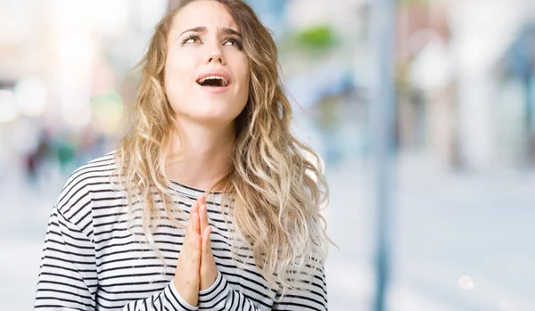 Beautiful young blonde woman wearing stripes sweater over isolated background begging and praying with hands together with hope expression on face very emotional and worried. Asking for forgiveness. Religion concept.