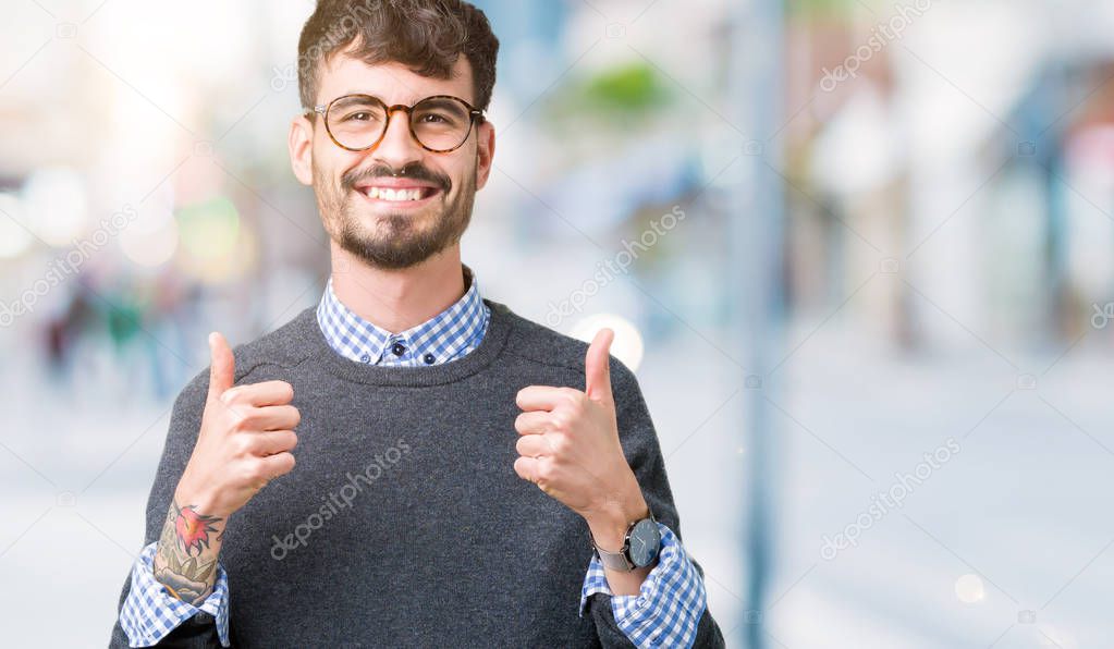 Young handsome smart man wearing glasses over isolated background success sign doing positive gesture with hand, thumbs up smiling and happy. Looking at the camera with cheerful expression, winner gesture.