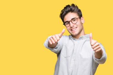 El ile pozitif jest yapıyor onaylama izole arka plan üzerinde gözlük takan genç yakışıklı adam başparmak gülümseyen ve başarı için mutlu. Kamera, kazanan jest arıyorsunuz.