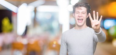 Gösterilen arka plan üzerinde kış kazak giyen genç yakışıklı adam izole ve parmakları ile işaret kendinden emin ve mutlu gülümseyerek süre dört numara.