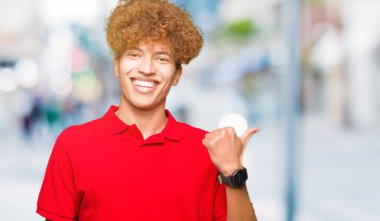 Kırmızı giyen afro saçlı genç yakışıklı adam mutlu ile gülümseyen t-shirt arıyorsunuz ve başparmak yanında işaret eden oktan yukarı yüz.