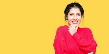 Kırmızı kazak ve emin çapraz kollar ve çeneme kaldırdı el ile gülümseme ile kameraya bakarak topuz giyen genç güzel kadın. Pozitif düşünce.