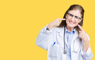 Orta yaş olgun doktor kadın kendinden emin gösterilen ve işaret parmakları diş ve ağız ile gülümseyen izole arka plan üzerinde tıbbi bir palto giyiyor. Sağlık kavramı.