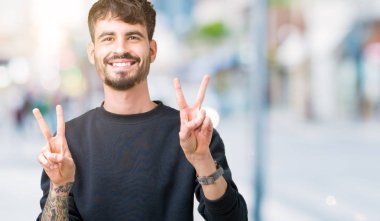 Arka planda kalan yakışıklı genç adam kameraya bakıp zafer işareti yaparken gülümsüyor. İki numara..