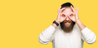 Dilini, parmaklarının arasından baktığını yapışmasını dürbün gibi ok jest yapıyor kış kazak giyen genç hipster adam. Deli ifade.