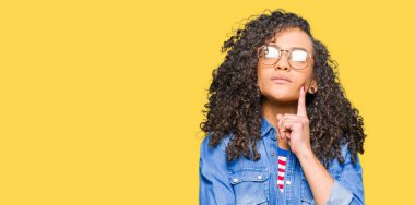 Kıvırcık saçlı gözlüklü el ile çene düşünme hakkında soru, dalgın ifade üzerine genç güzel kadın. Düşünceli yüzüyle gülümsüyor. Şüphe kavramı.